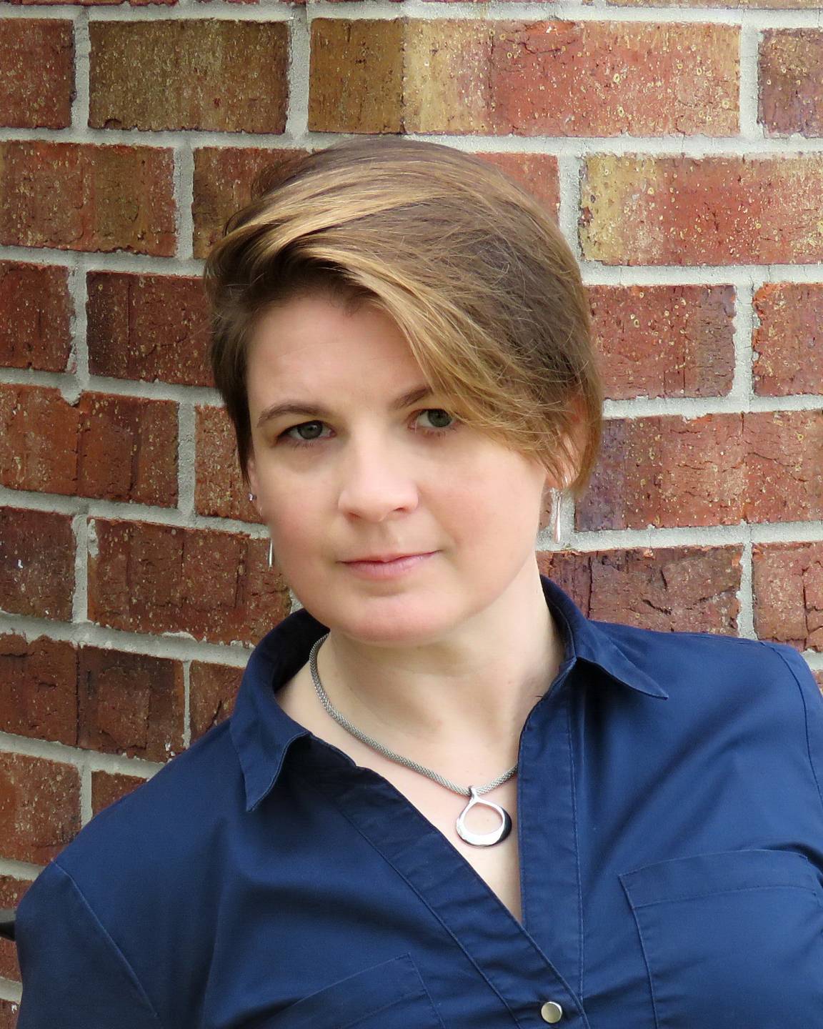 Jennifer Brooks has short brown hair and is wearing a blue button up shirt. There is a brick background