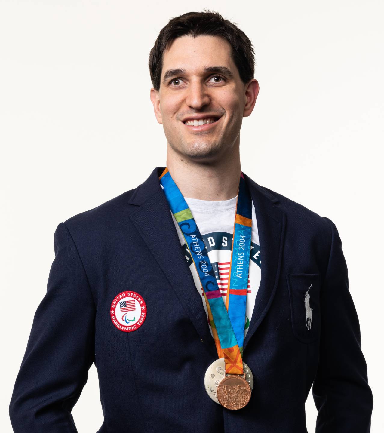 Tyler Merren has short dark hair and is wearing a dark blazer with a white Team USA shirt underneath. There are several medals hanging around his neck