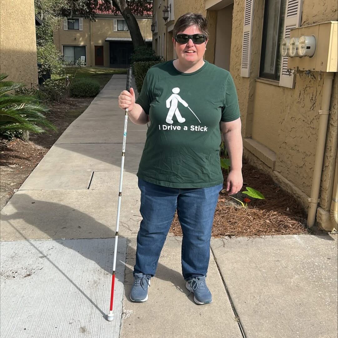 Alexis stands on the sidewalk near her home with her cane in hand. She is wearing a shirt that says "I drive a stick" that has an image of a person walking with a cane.