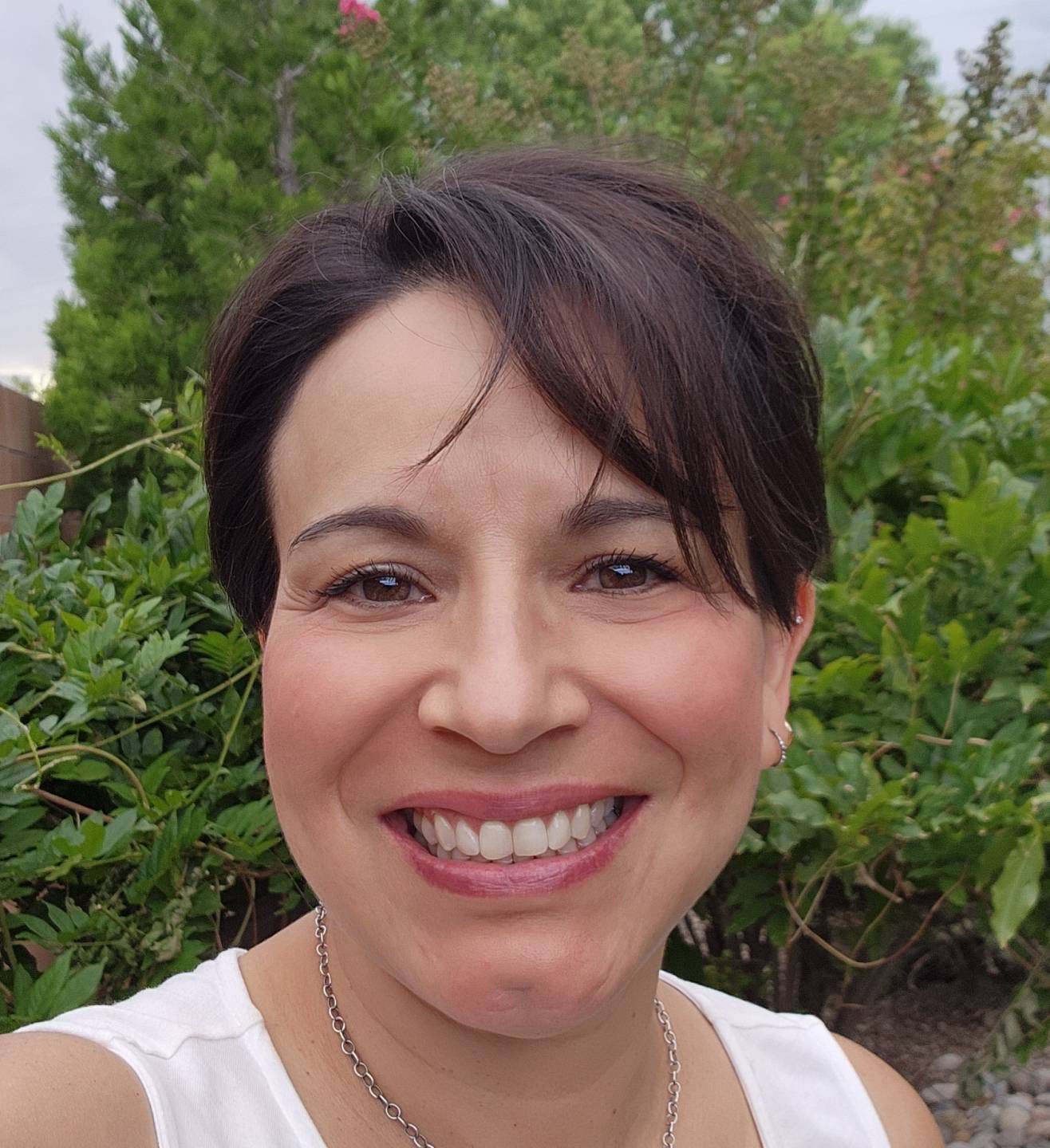 Andrea Montano has short dark hair and is wearing a white sleeveless top. There is greenery in the background.