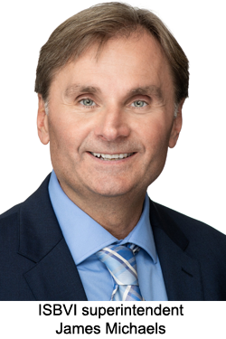 James Michael has short light brown hair and is wearing a blue button up shirt and blue tie with a dark jacket