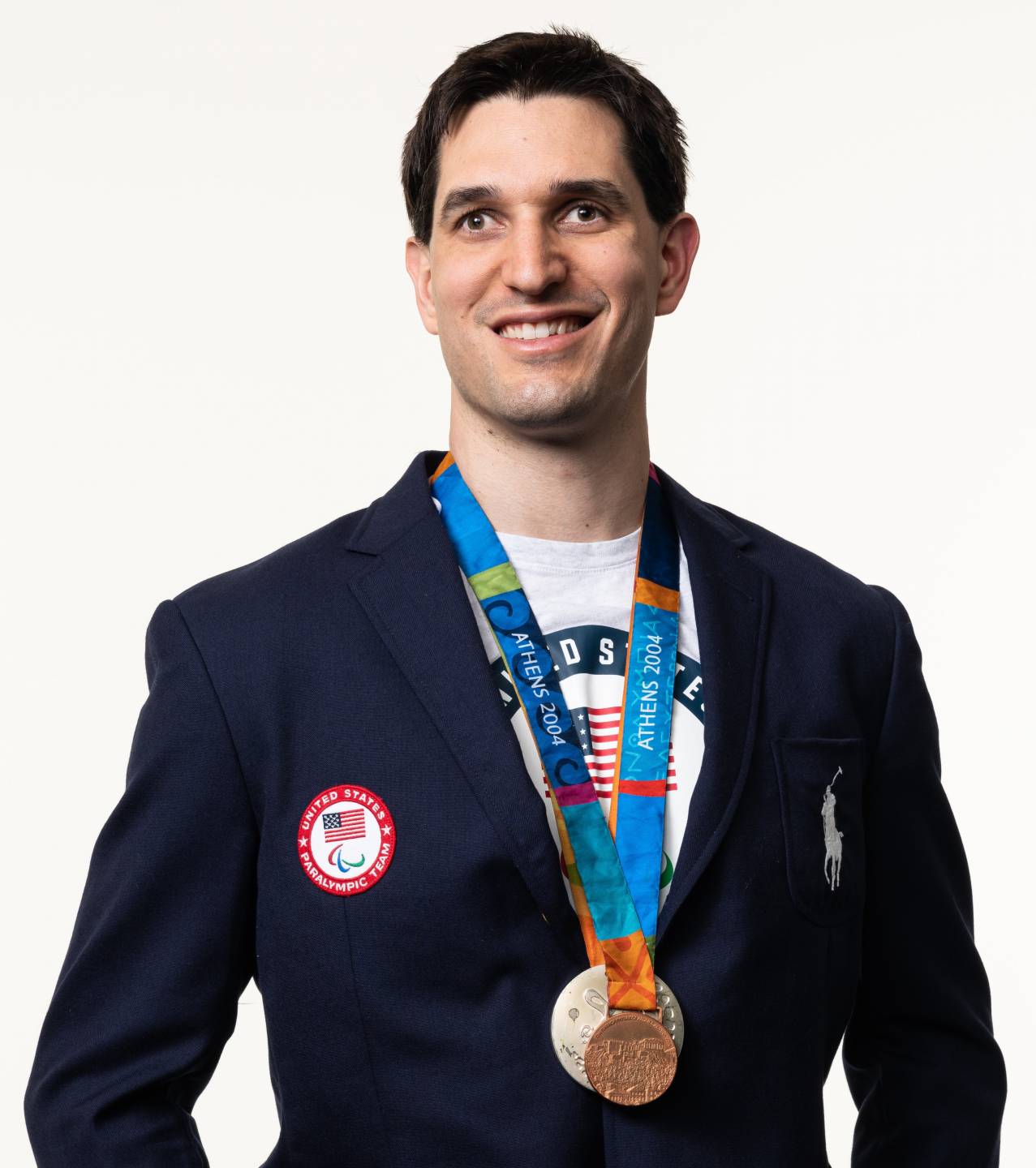Tyler Merren has short dark hair and is wearing a dark blazer with a white Team USA shirt underneath. There are several medals hanging around his neck,