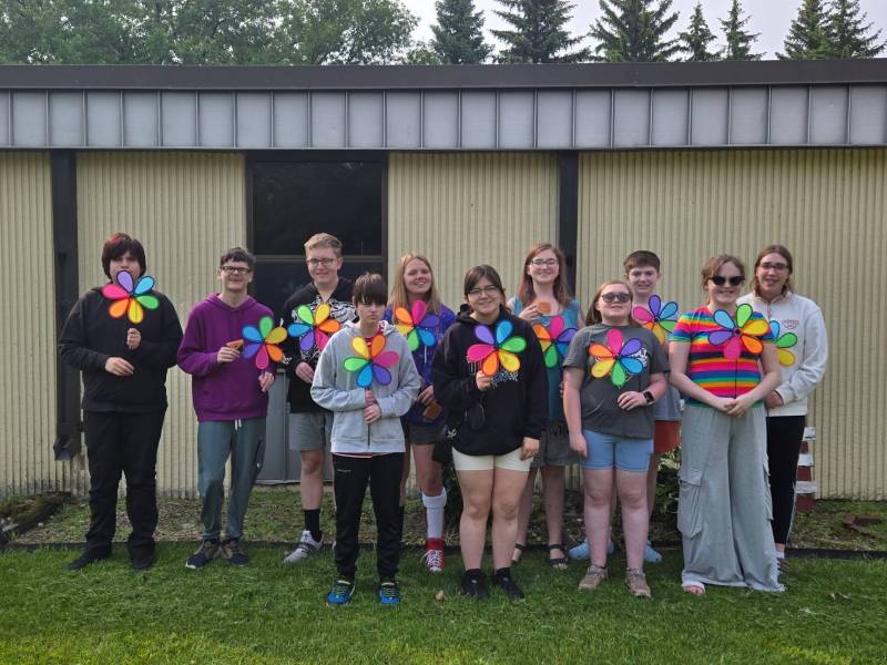 11 teens stand in two rows outside of a building. They each hold a colorful lawn spinner. Most are smiling.