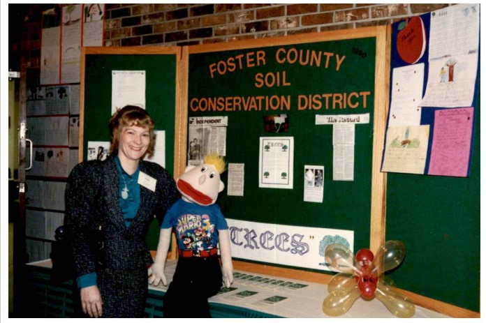 Nedra smiles next to a bulletin board holding a life-size puppet with a wide mouth, spiky blond hair and a blue t-shirt on.