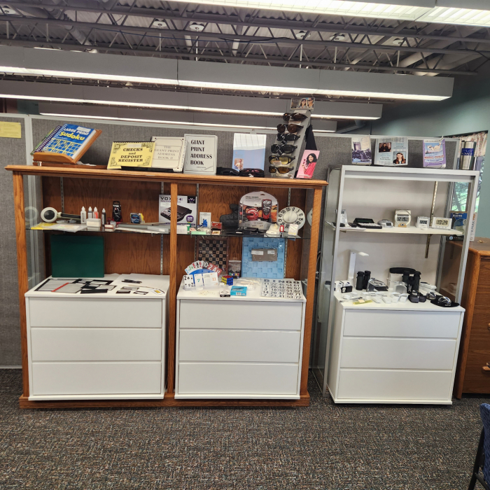 3 shelving units stand next to each other full of Store products. The bottom portion has drawers and the top portion are open shelves. There are a variety of products, including magnifiers, talking clocks, large print books, tactile glue, writing guides, and many other assistive tech devices shown.