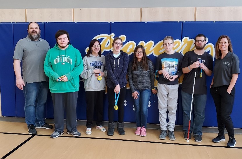 6 students and 2 staff members smile and stand in a line in the gym at South Dakota's gym where they played goalball.