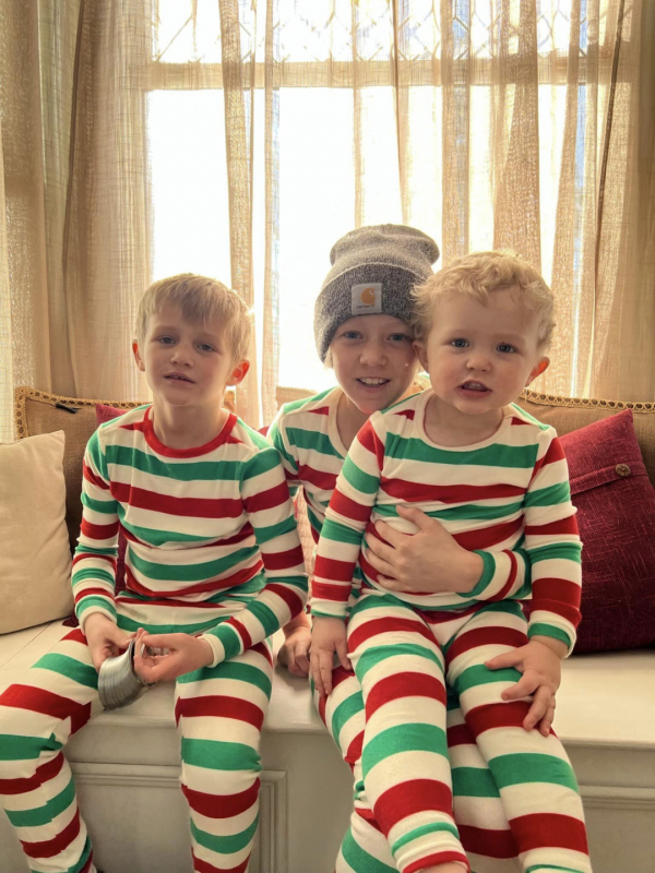 3 young boys sit on a bench in front of a window wearing matching striped Christmas pajamas. One holds a slinky. The youngest boy sits on one of the older boy's lap. They smile at the camera.