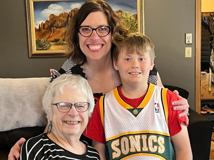 A close-up of Carmen (seated) with Amanda standing behind her and Carter standing to her left. Amanda has her arms draped around each of them. They are all smiling at the camera.