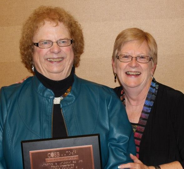 Carmen and Marje stand next to each other smiling at the camera. Carmen is holding a plaque in front of her and Marje's left hand holds onto Carmen's arm.