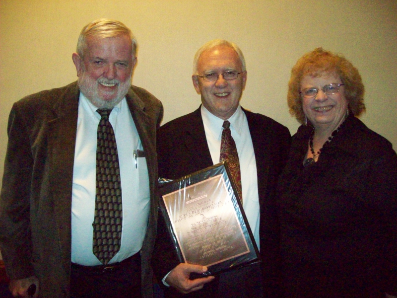 Carmen stands on the right side of a group of 3. Dr. Phil Hatlen stands on the left and Dr. Ralph Bartley, who is holding an award, stands in the middle. All are wearing dress clothes and are smiling at the camera.