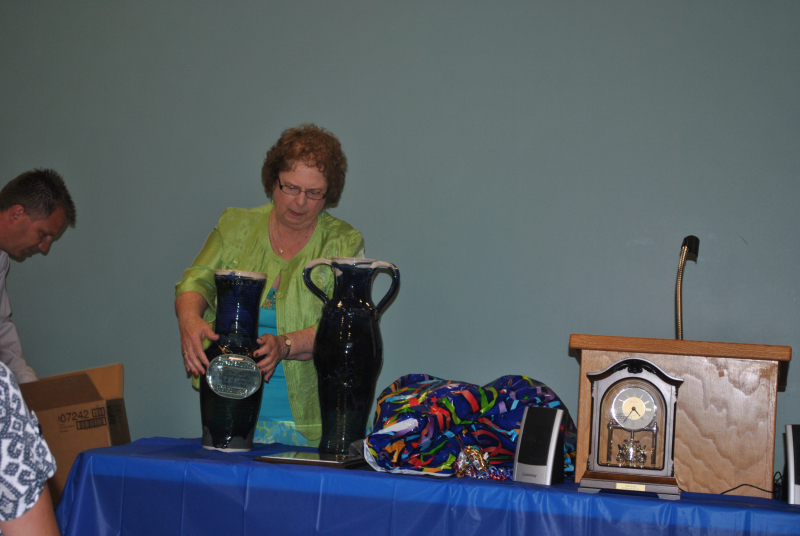 Carmen receiving the two blue clay vases at her retirement party in 2013. She is standing behind a table with her hands around one of them to pick it up.