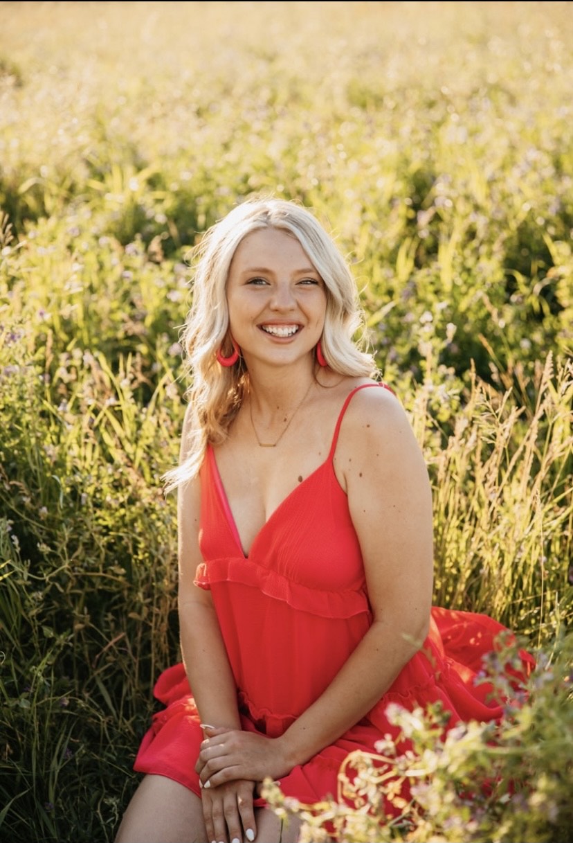 Cylee sits in a field of grass wearing a red dress. She has long wavy blond hair and is smiling at the camera.