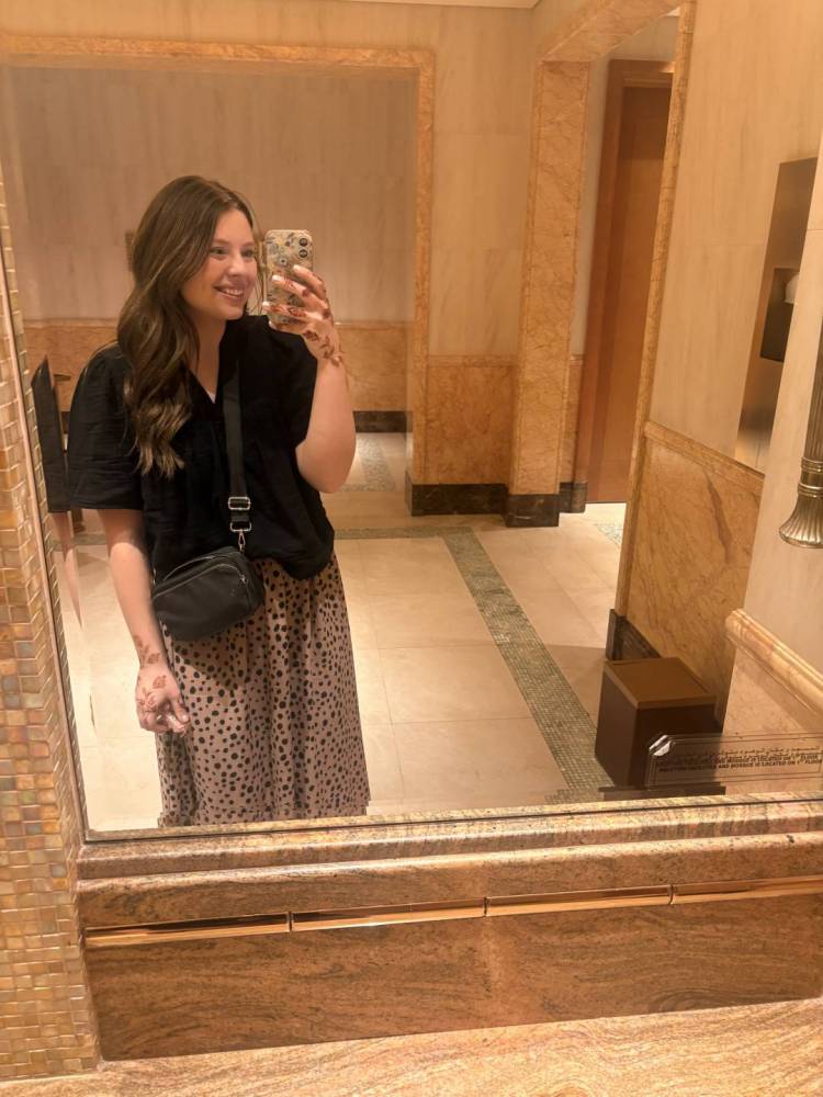 Cylee takes a selfie with her phone as she stands in front of a mirror. She has long light brown hair and wears a black blouse and an animal print skirt.