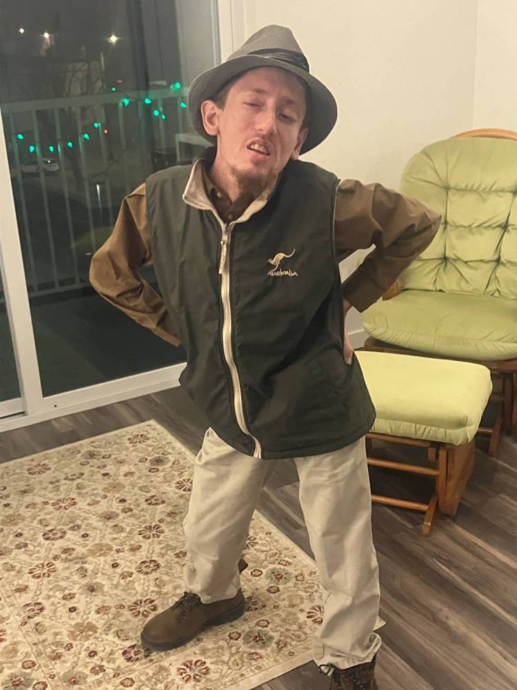 Wearing a hat, a brown shirt, a black vest, and khaki pants, Dietrich poses in his apartment.