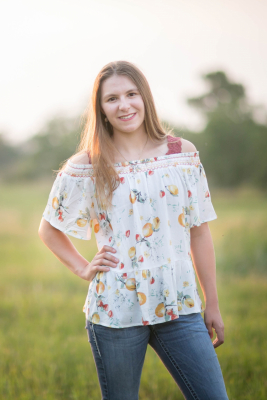 Faith stands in a field wearing a white floral top and blue jeans.