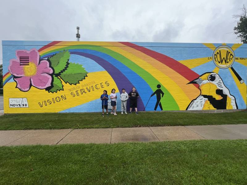 4 elementary students stand in front of a largem, colorful mural on the side of a building.