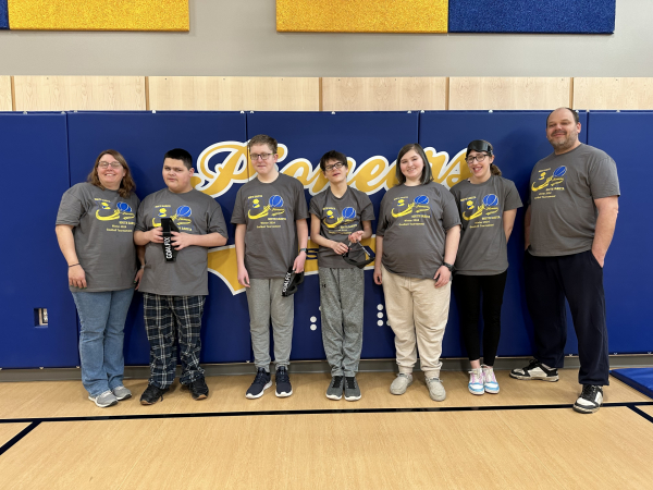 5 students and 2 adults wearing goalball tees stand in a line against a wall in a gym.