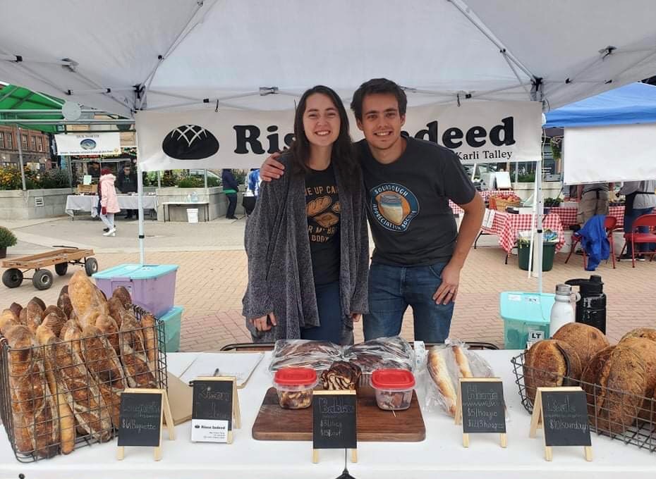Karli and her husband stand close together. He has his arm around her shoulders. They are standing under a tent and behind a table with many different loaves of bread on it.