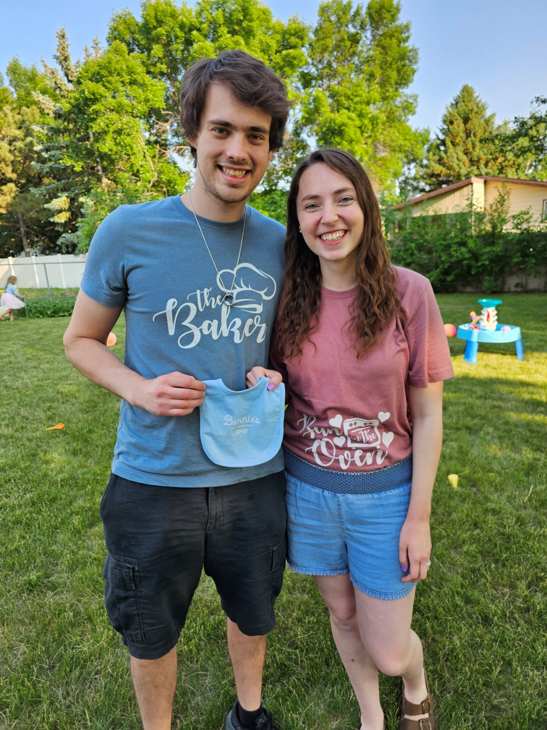 Karli stands next to her husband. He is holding a blue bib and wearing a shirt that says "the baker." Karli's shirt says "bun in the oven."