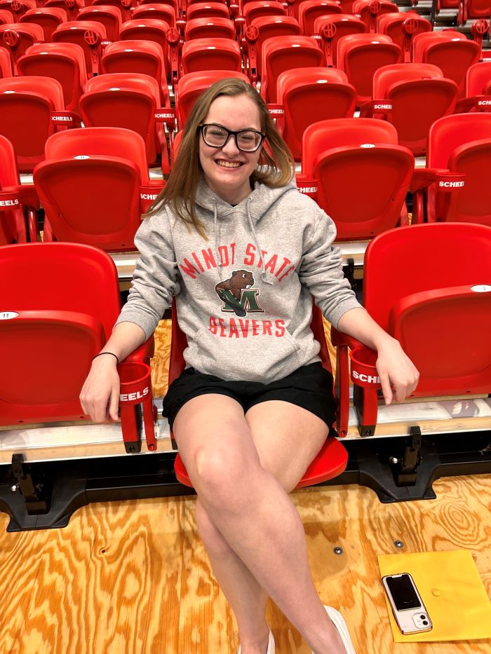 Wearing a Minot State Beavers sweatshirt, Kaylyn smiles as she sits in a bleacher chair in the auditorium at Minot State University.
