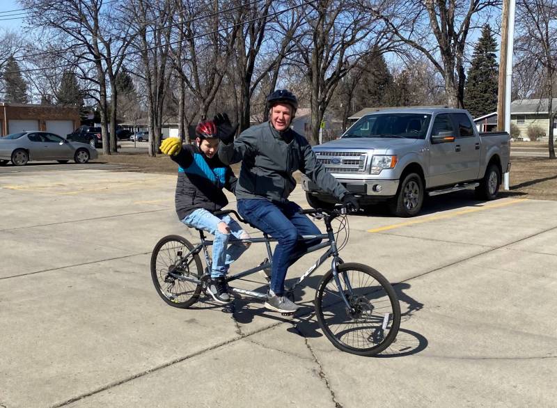 Two men ride on a tandem bike in a parking lot. Both wear gloves, helmets, and winter coats.