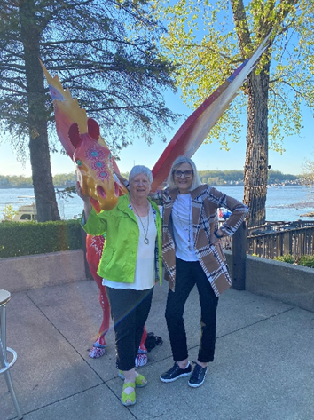 Marje and Lanna stand next to each other outside in front of a statue of a red and yellow horse. 