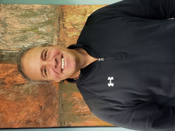 Jody, who has gray hair and is wearing a black sweatshirt, stands in front of a orange and brown abstract painting smiling.