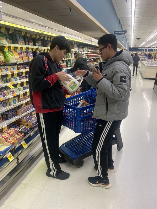 Matthew and a fellow student stand in the dairy aisle of the grocery store. Matthew holds a gallon of milk up to the other boy while they stand in front of a cart.