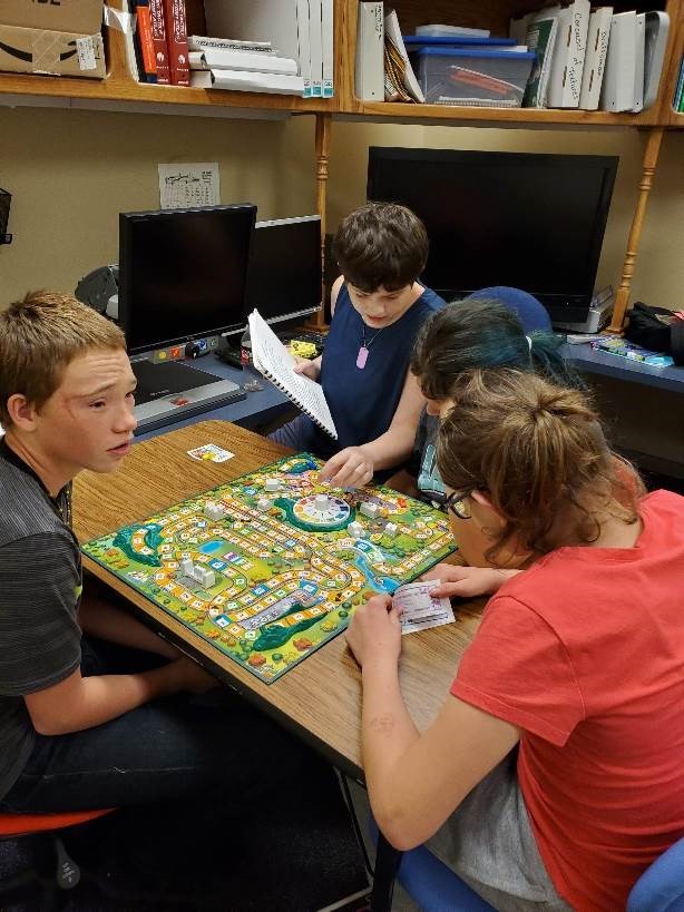 3 students sit at a table playing the game of LIFE. One student reads the directions to the others.