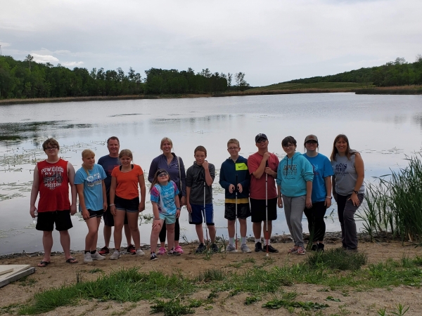 Middle Schoolers and 2 adults from Annie's House stand on a beach in front of a lake.