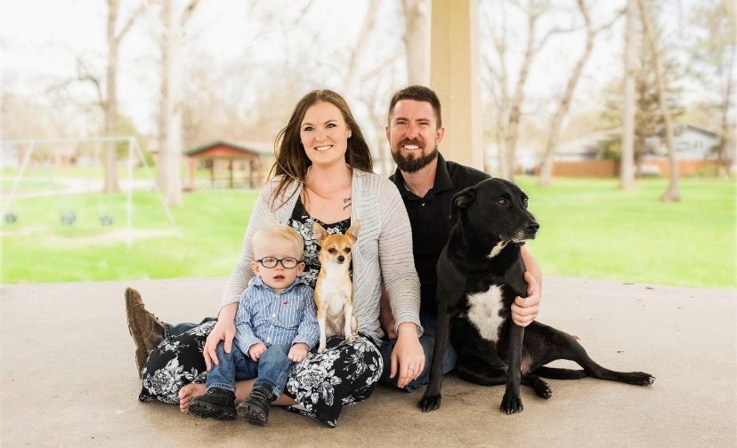 Morgan sits on the ground at a park smiling with her boyfriend Jeddrey and son and their 2 dogs.