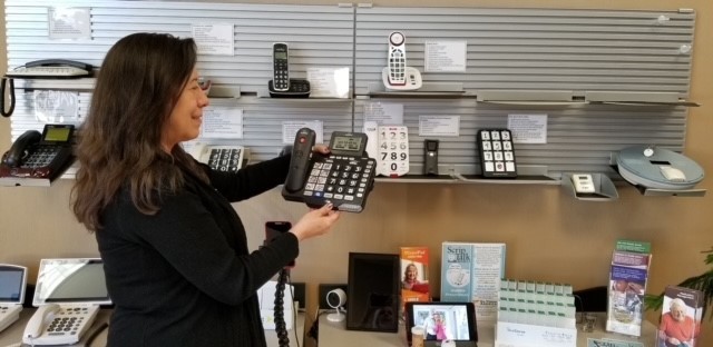 A woman stands in front of shelves with a variety of phones. She is looking at, smiling, and holding one with larger buttons and a display at the top. 