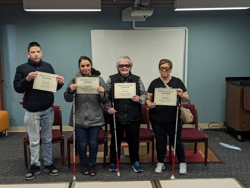 4 adults proudly hold their certificates of courage in front of them while standing in the library at NDVS/SB