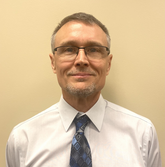 Wearing a white dress shirt and blue and gray checkered tie, Paul Olson smiles at the camera