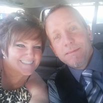 Paula and Arik sit in a car and lean towards each other as they take a selfie. They are wearing dress clothes and are smiling at the camera.