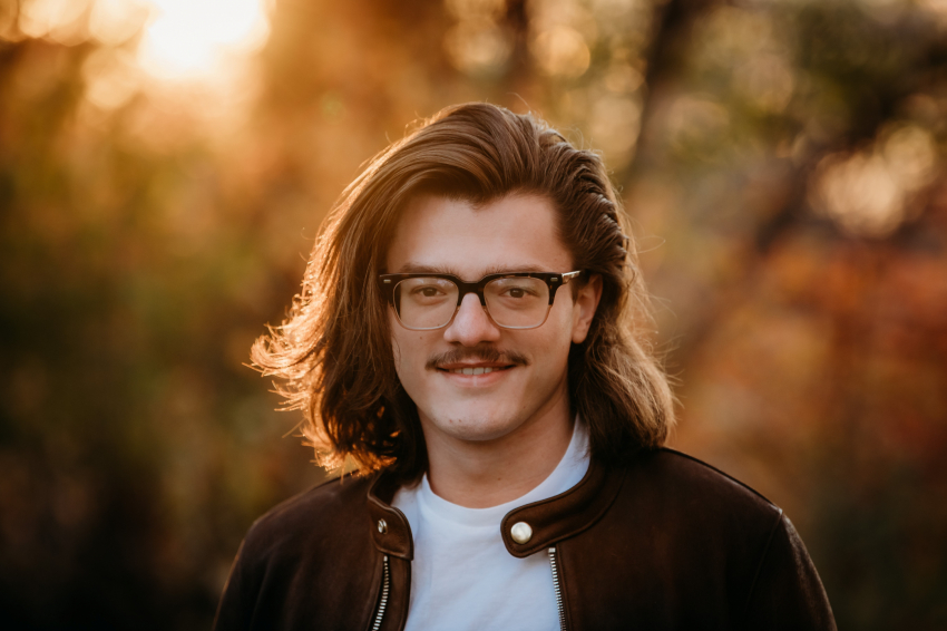 Paxton stands outside wearing glasses, a brown suede jacket and a white t-shirt.