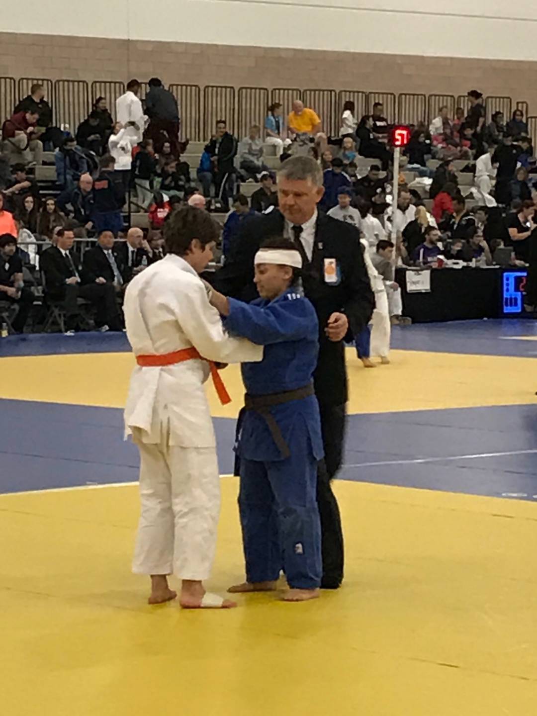 Quincie meets her opponent on the judo mat while a judge looks on. She and her competitor are touching each other's sleeves before the match begins.