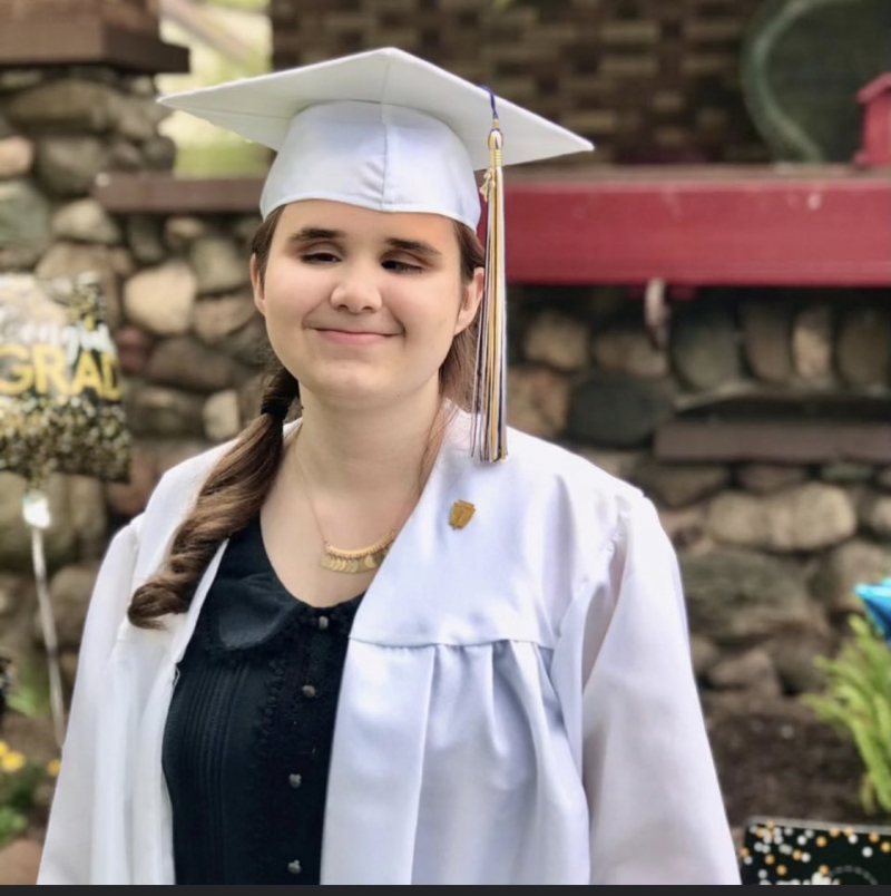 Rowan stands in their white graduation cap and gown. Their brown hair is tied in a ponytail and they are wearing a black button-down shirt and gold necklace underneath the gown.