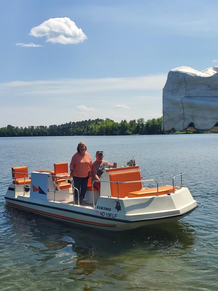 Tami stands on a boat next to her husband Brian who is steering the boat, which is on a lake. 