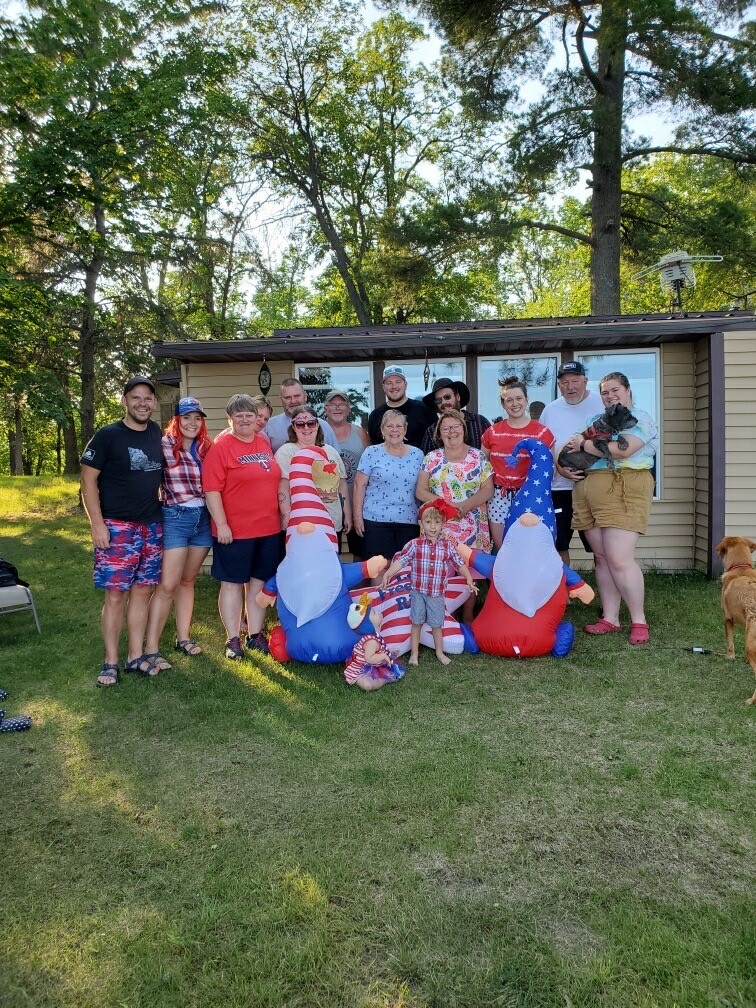 Tami stands with her extended family in front of her cabin on the 4th of July. Everyone is smiling and wearing summery clothing.