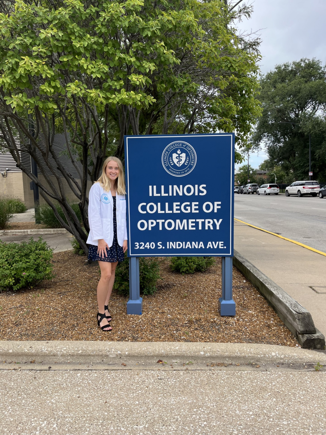 Abby stands by a sign that reads Illinois College of Optometry while wearing a white coat representing her work towards her doctorate degree. She smiles at the camera.