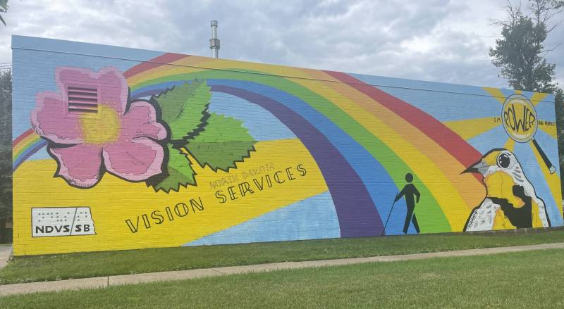 A mural with a prairie rose, a meadlowlark, a rainbow, a figure walking with a cane, and the words North Dakota Vision Services empowering people graces the side of a brick building.