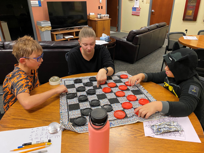 Amy sits at a table in the Commons at NDVS/SB between 2 elementary age boys playing checkers.