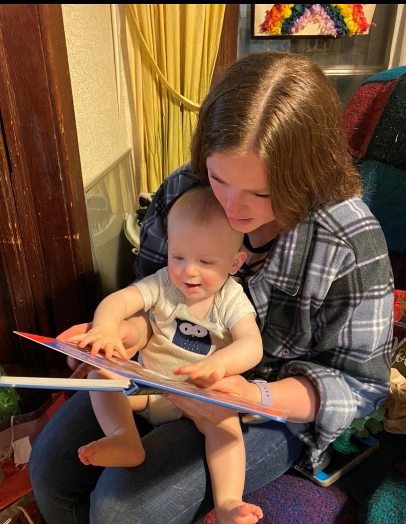 Morgan sits in a chair holding her son while reading a book to him. He is reaching for the pages.