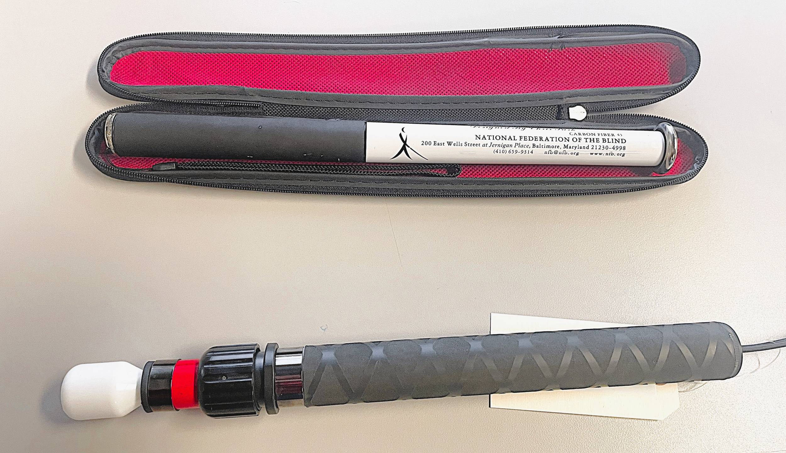 canes that collapse into themselves instead of folding. The NFB7 cane is all white with a black handle and a flat metal tip. The Ambutech cane is red and white with a black handle and a marshmallow hook tip.