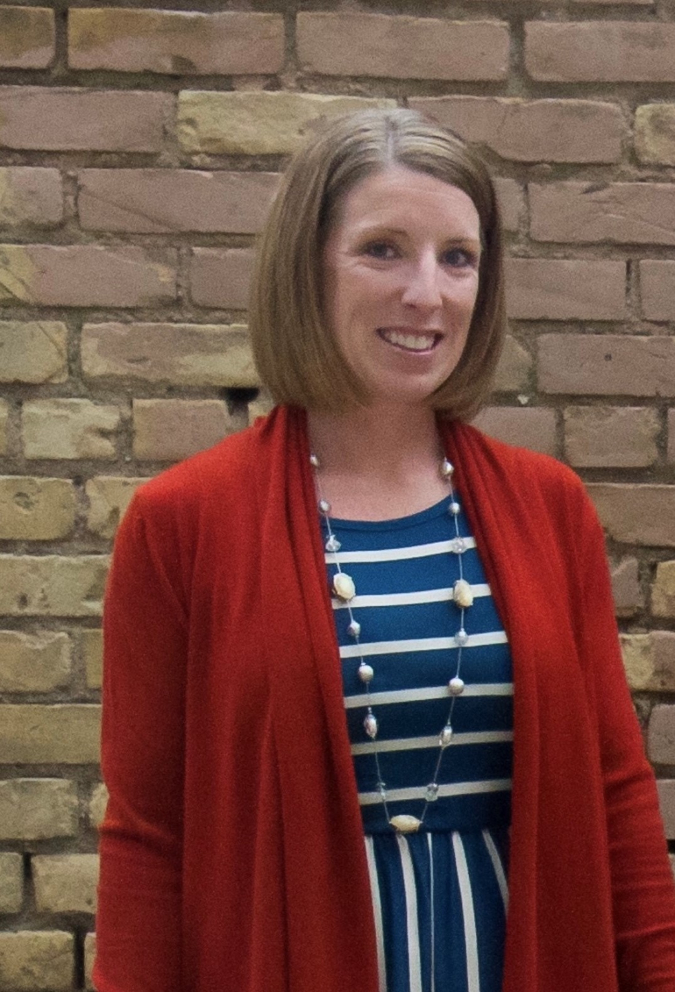 Danielle stands smiling in front of a brick wall wearing a blue and white striped dress, a red cardigan and a long necklace. She has chin-length brown hair.
