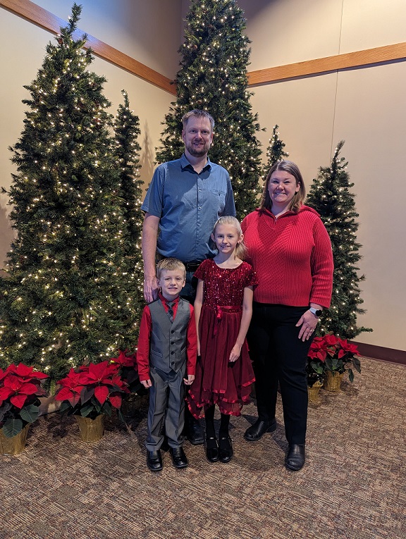 David stands in front of a group of Christmas trees with his wife and their young son and daughter. They are all wearing dress clothes and smiles.