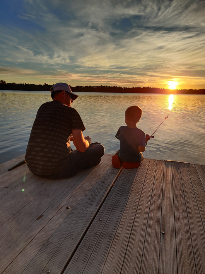 At sunset, David sits on a dock next to his young son who is holding a fishing line off the dock into the water. 