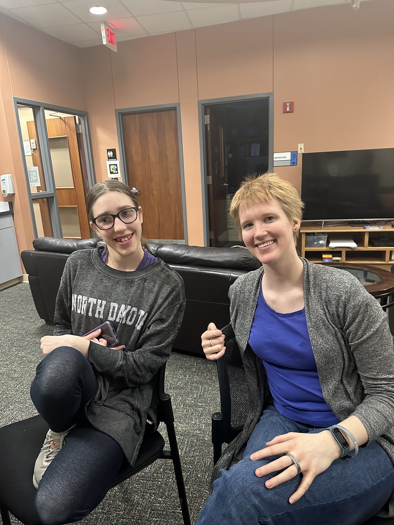 Gretchen, who has short red hair and is wearing a blue t-shirt with a black cardigan sits next to a middle school aged girl in the Commons. They are smiling at the camera.