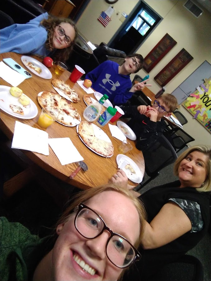 Gretchen's face is at the bottom of the image as she is taking the selfie. Three students and another houseparent are seated at the table behind her with pizza, plates, and other dinner necessities.